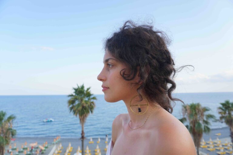 ragazza di profilo, con capelli scuri mossi e un vistoso orecchino dorato circolare. Lo sfondo della foto ritrae una spiaggia assolata con palme e ombrelloni vicino al mare.